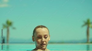 Beautiful young woman with blonde hair in brown swimsuit coming out from under pool and smiling at camera. Blonde-haired woman enjoying the pool and vacation. The camera is at the pool water level.