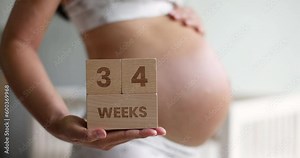Pregnant woman showing 34 weeks pregnancy bump for maternity photoshoot holding wooden blocks sign happy. Asian beautiful model happy standing at home.