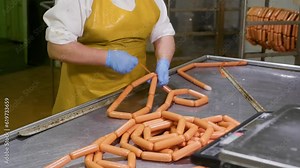 Butcher woman processing sausages tying knots at meat factory on conveyor line. Worker female makes sausage products on automated production equipment at meat plant. Food manufacturing, meat industry.