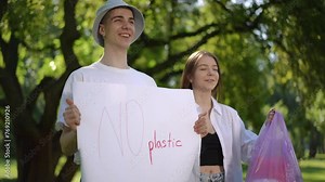Protest say no to plastic. A guy and a girl stand motionless on a summer day outdoors in a city park. The guy is holding a No plastic poster in his hands, and the girl is holding a plastic bag with