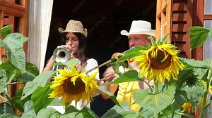 A young woman and a man play the trumpet and trombone in a country house by an open window overlooking the flowers in the garden.Active musicians in hats blow into a musical instrument in the sunlight