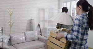 slow action of two asian sisters walking in new apartment with moving carton boxes put down heap. smiling happy girls friends outstretched arms relax finally finish relocation parcels talking laugh