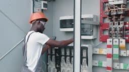 Man in uniform repairing cables in switchgear at solar farm. African...