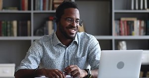 Smiling african male student wear headset elearning study with online tutor teacher talk conference calling do video chat learn language make notes sit at home office desk look at laptop computer