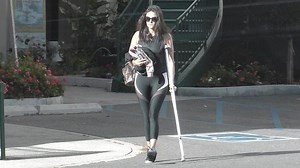 Nina Dobrev hobbles to her car, hands full, using a crutch