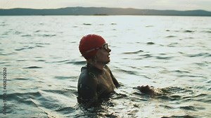 Female triathlete in wetsuit and cap standing in lake and taking off goggles after swimming workout