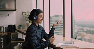 Young confident Indian business lady sit in office kitchen at bar counter wear headphones talk to company client remotely use laptop video conference call app, communication by modern tech concept