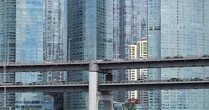 Slow-moving traffic fills elevated viaduct at end of bridge, set against backdrop of modern glass-walled skyscrapers. Rush hour congestion sees cars moving slowly along busy lanes in both directions.