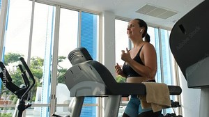 Cheerful young woman doing cardio training on treadmill, slow motion. Female listening to music in headphones while working out in a gym