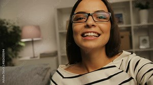 Close up shot of attractive happy woman in glasses using camera for video chatting with friends, family at home in the living room at the evening.