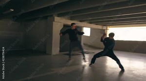 Wide angle fight scene in a car park. Men in dark clothes battle in hand to hand combat in an urban setting. Shadowy parking garage with men fighting near parked cars.