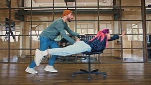 Office superhero. Young man riding his colleague woman with pink hair on chair pretending she is flying, side view