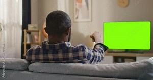 African American man sitting at sofa at modern appartment. Male holding remote control and changing channel. Man looking at chroma key TV set. Male sitting at couch and looking at green moke up TV.