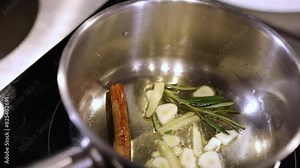 Garlic simmering, Oil infusion, Aromatic herbs. Close-up sliced garlic, rosemary, and cinnamon stick simmering in oil in stainless steel pan.
