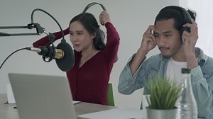 woman was speaking on a morning radio station broadcasting a news program and men preparing the content via computers. The woman is testing the microphone by speaking test. concept podcast .