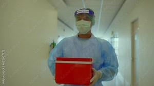 A healthcare worker in full protective gear transports a red container with medical samples through a hospital corridor, ensuring safety and proper handling.
