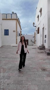 Woman cheerily walks in an alley between white houses. She is enjoying the atmosphere in Rabat, Morocco. She turns her face, smiles, and raises her arms to the camera.