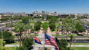 American flag, Arizona state flag, and POW MIA flags waving in on capitol grounds. Aerial rising shot revealing Phoenix, Arizona skyline.