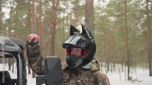 Two riders with helmets beside atv in snowy forest, driver and guide check windshield and engine on camo utility buggy, breath visible in cold air, pine trees and soft falling snow create tense