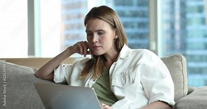 Serious thoughtful freelance woman watching video presentation on laptop, using computer at home, sitting on couch, thinking over content, giving reaction, getting happy, smiling, laughing