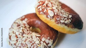 Two donuts coated with milk chocolate glaze and sprinkled with nuts rotate on a platform on a white background.