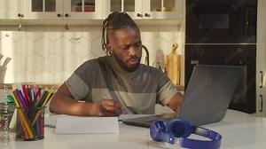 Focused serious handsome African man with laptop pc and notepad calculating finance, planning family budget and payment, shocked by bills, expressing worry and frustration in domestic kitchen.