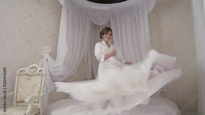 A happy bride with makeup and hairstyle in a lacy dressing gone jumps on the bed holding her glamorous wedding dress. Inside shooting, white baldachin over the bed is on the background.