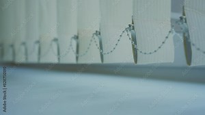 Close up of white vertical blinds with connecting chain at bottom. Soft light creating gentle shadows on surface.