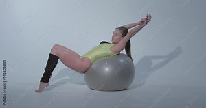 Beautiful girl in a yellow sports suit and black leggings is engaged in gymnastics on a gray rubber ball, swimsuit, beautiful skin, figure, barefoot, crouchs on the ball, jumps, slog