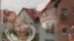 Elderly married couple standing by window together, caring husband hugging wife. Couple in nursing home during lockdown, loneliness and depression concept