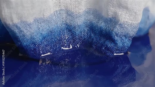 White fabric slowly dipping into blue liquid in slow motion. Macro close-up of absorption and dyeing process. Textile and chemistry concept