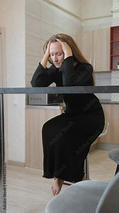Young woman sitting on bar stool in kitchen, holding her head in her hands, struggling with headache. Frustration and exhaustion are evident as she deals with her pain. vertical video