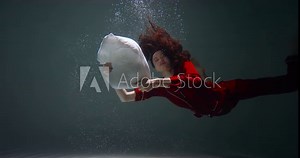 Cinematic shot, young beautiful mixed race woman in red pyjamas sinks under water to fall asleep with pillow slow motion
