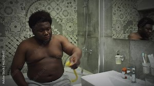 Overweight Black man with shirtless torso sitting on edge of bathtub, taking measurements of waist with tape measure and feeling sad