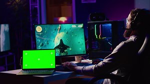 Green screen laptop next to man using gaming keyboard to play spaceship flying singleplayer game. Mockup notebook and gamer navigating with spacecraft in galaxy, shooting at asteroids
