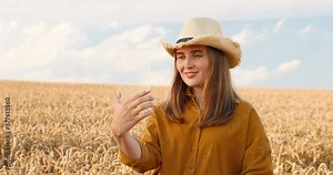 Portrait of beautiful happy Caucasian girl standing in golden field and using futuristic gadget outdoor. Pretty young woman in hat tapping on holographic device in countryside. Technology concept