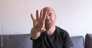 bald man with a red goatee sitting on the couch at home. Wearing a black shirt, he counts down from 5 to 0 and celebrates excitedly.