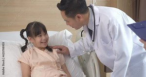 Asian male doctor and female nurse with a clipboard standing talking with a smile and consoling at bedside with a little sick girl in patient room at hospital. Medical Healthcare concept.
