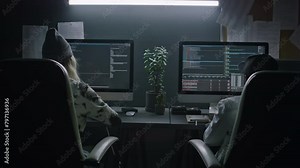 Medium rear of two polyethnic software developers sitting in chairs at desks in office at nighttime, writing computer code, testing for errors and variables, while working together on project