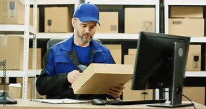 Caucasian postman in uniform working at computer in post office store. Postman scanning bar code with scanner, registering parcel and filling in invoice document while sitting at desk. Stock Video