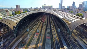 Aerial view of the station. Trains arrive at the station. An old wide arched structure of metal and glass above the station pillars. Tourism. Skyline with tall buildings. Italy, Milan, June 2022