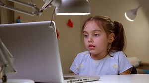 Cute preschool girl is sitting at home at a desk and at a laptop, talking on a video call. Online e-learning class, studying with a teacher, social distance learning, home learning concept.