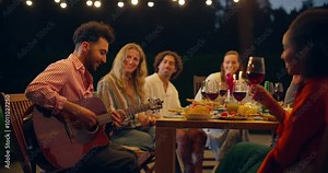 Fun Night Gathering of Diverse Friends Around a Table Under String Lights. One Man Entertaining Everyone by Playing Guitar. Others Dance, Sing and Enjoy the Music, Adding to the Lively Mood