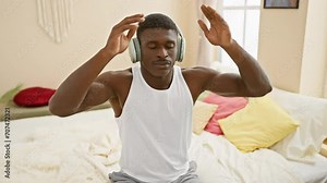 An adult black man practices yoga at home in a bedroom while listening to music on headphones, illustrating a peaceful atmosphere.