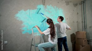 Rear view two painters paint wall together with rollers. Girl standing on building ladder. Home renovation, boxes and concrete walls. Married couple working, covering walls with primer or blue paint.