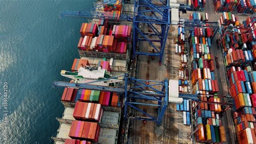 Top-down aerial drone view of port cranes loading and unloading shipping containers at a busy cargo terminal. Industrial logistics, global trade, maritime transport and supply chain infrastructure con