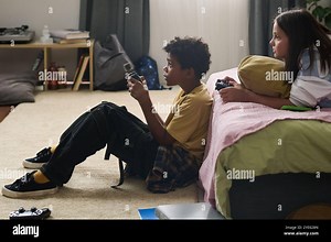 Two friends sitting on floor in a cozy bedroom, intensely playing video games together, surrounded by gaming devices and bookshelves. Scene exudes fun and camaraderie Stock Photo - Alamy
