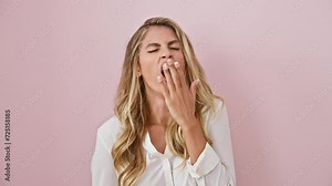 Beautiful blonde woman, overwhelmed by sleepiness, exhibiting a huge yawn. standing bored and tired over an isolated pink background, her hand does its best to cover her yawning mouth.