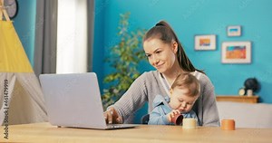 Young smiling mom sitting at desk in front of the computer screen, working remotely on laptop, writing messages, holding her little son on lap playing with toys, child is polite
