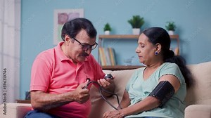 Indian senior man checking blood pressure or bp of wife by using sphygmomanometer while sitting on sofa at home - concept of home health checkup, relationship and caring husband.